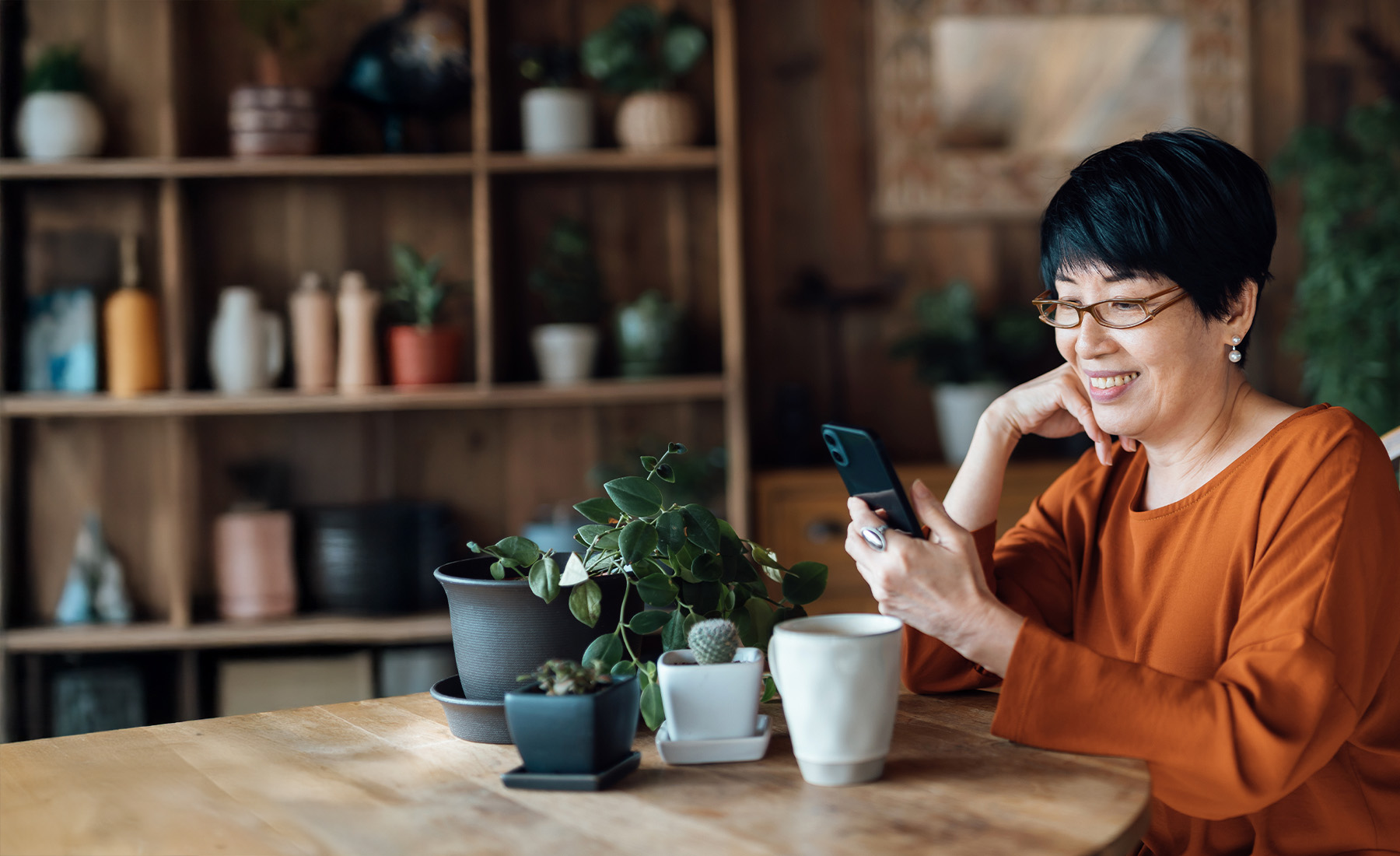 A woman wearing glasses talks on the phone at home. She sits at a table with a selection of potted plants.